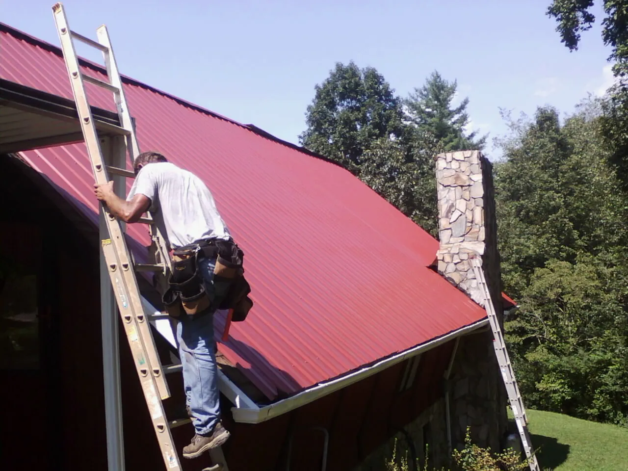 Roofer on a ladder beside a red metal roof and stone chimney — Soffit & Fascia Repair by Roofing Brothers in Cornwall
